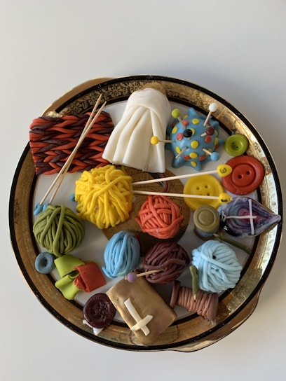 A decorative platter containing an assortment of handcrafted items resembling yarn balls, a white draped cloth, buttons, and a spherical blue object with colorful pins. These items are creatively arranged on an ornate golden dish.