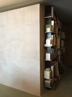 A minimalist bookshelf is built into the side of a wall. The shelves are populated with various books and documents, organized neatly. The wall is painted in a light, neutral color, which contrasts with the bookshelf's dark finish. The overall room appears to have a modern and simplistic design.