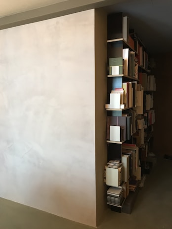 A minimalist bookshelf is built into the side of a wall. The shelves are populated with various books and documents, organized neatly. The wall is painted in a light, neutral color, which contrasts with the bookshelf's dark finish. The overall room appears to have a modern and simplistic design.