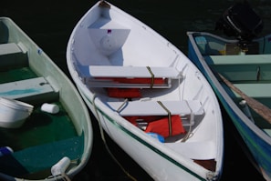 A white rowboat is docked alongside two other boats. It contains several red life jackets and a rope coiled inside. The boat to the left is pale green with a few objects floating inside, possibly plastic containers. The boat to the right is light blue with an outboard motor attached.