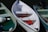 A white rowboat is docked alongside two other boats. It contains several red life jackets and a rope coiled inside. The boat to the left is pale green with a few objects floating inside, possibly plastic containers. The boat to the right is light blue with an outboard motor attached.