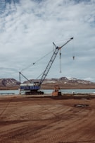 A large blue crane stands on a construction site near a body of water with mountainous terrain in the background. The ground appears rough and industrial with some construction materials scattered around.