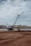 A large blue crane stands on a construction site near a body of water with mountainous terrain in the background. The ground appears rough and industrial with some construction materials scattered around.