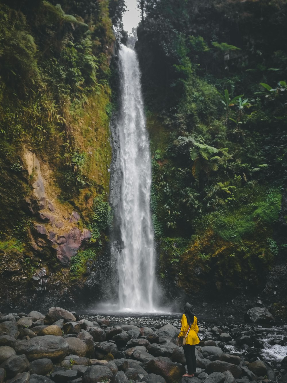 person in yellow jacket standing on rocky shore near waterfalls during daytime