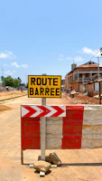 A construction site with a 'Route Barrée' (Road Closed) sign in the foreground. The sign is yellow with black text and stands on a makeshift barricade with a red and white striped design. In the background, there's a dirt road under construction, buildings to the side, and a clear blue sky.