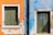 Two side-by-side wooden shutters are mounted on a weathered, textured wall. The wall is divided into two sections, with the left side painted in a bright orange and the right side in a faded blue. The shutters are green and appear aged, with signs of wear and peeling paint. There is a vertical black pipe running between the two sections of the wall.