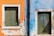Two side-by-side wooden shutters are mounted on a weathered, textured wall. The wall is divided into two sections, with the left side painted in a bright orange and the right side in a faded blue. The shutters are green and appear aged, with signs of wear and peeling paint. There is a vertical black pipe running between the two sections of the wall.