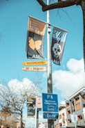 Colorful signs and banners are attached to a post against a clear blue sky. Two large museum banners display artwork and promotional text. Street signs beneath point to accommodations and provide parking information. A leafless tree and urban buildings are visible in the background.