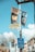 Colorful signs and banners are attached to a post against a clear blue sky. Two large museum banners display artwork and promotional text. Street signs beneath point to accommodations and provide parking information. A leafless tree and urban buildings are visible in the background.