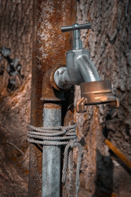 A metal water tap is attached to a rusty pipe, with a rope tied around the pipe. The background consists of a textured tree bark, providing a rustic and natural setting.