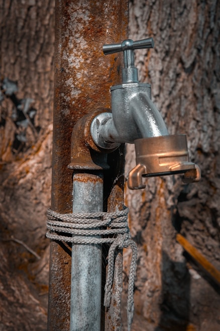 A metal water tap is attached to a rusty pipe, with a rope tied around the pipe. The background consists of a textured tree bark, providing a rustic and natural setting.