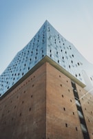 A modern architectural building with a sleek glass facade and a contrasting brick base. The glass section features reflective windows with a wavy design pattern, creating a visually striking contrast against the rigid structure of the brick.