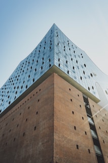 A modern architectural building with a sleek glass facade and a contrasting brick base. The glass section features reflective windows with a wavy design pattern, creating a visually striking contrast against the rigid structure of the brick.