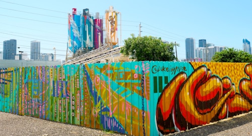 A vibrant mural covers a long wooden fence with an urban background featuring industrial silos painted with artistic designs and modern city skyscrapers in the distance. The mural showcases a variety of colorful patterns, shapes, and abstract forms in bright hues.