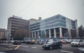 A modern commercial building with a grid of large blue-tinted windows beside an older, more traditional structure with a company logo. Cars are parked and in motion around the area, and the streets are lined with road signs and traffic lights. Leafless trees are scattered around the perimeter, indicating a possible winter setting.