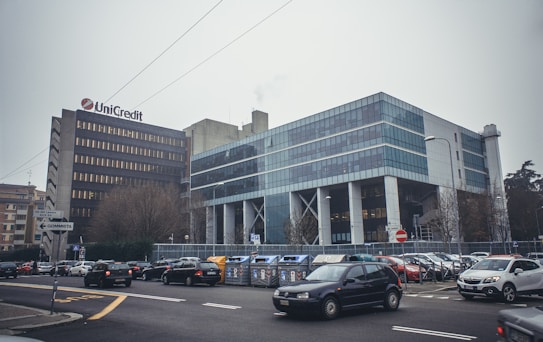 A modern commercial building with a grid of large blue-tinted windows beside an older, more traditional structure with a company logo. Cars are parked and in motion around the area, and the streets are lined with road signs and traffic lights. Leafless trees are scattered around the perimeter, indicating a possible winter setting.