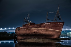 A large rusted ship labeled N182 is stranded on a muddy shore at night. The dimly lit bridge in the background casts a faint glow over the scene, reflecting on the water. The sky is dark and overcast, adding to the eerie atmosphere.