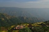 A small village with a cluster of houses is situated in a valley surrounded by lush green mountains and terraced fields. The sky above is partly cloudy with a light haze covering parts of the mountains in the background.