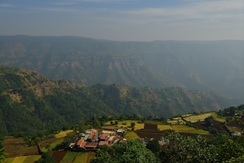 A small village with a cluster of houses is situated in a valley surrounded by lush green mountains and terraced fields. The sky above is partly cloudy with a light haze covering parts of the mountains in the background.