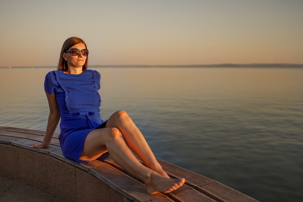 woman in blue dress sitting on brown wooden dock during daytime