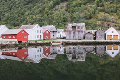 A row of colorful wooden houses is reflected perfectly in the calm water of a fjord. The houses are set against a lush, green hillside covered in dense trees and foliage. A single boat floats near the shore, enhancing the serene atmosphere. The buildings range in color from red to white and gray, showcasing traditional Scandinavian architecture.