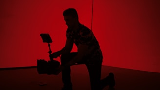 A silhouette of a person kneeling is holding a camera on a stabilizer against a vivid red background.