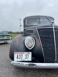 A vintage black truck with Wisconsin license plate A37 37 displayed on a wet pavement. The truck features a large round headlight and a prominent grille design. Another vehicle, possibly another classic car, is partially visible in the background, set against a cloudy sky and a grassy field.