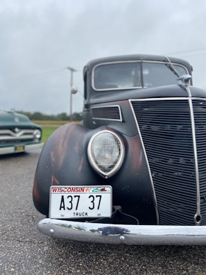 A vintage black truck with Wisconsin license plate A37 37 displayed on a wet pavement. The truck features a large round headlight and a prominent grille design. Another vehicle, possibly another classic car, is partially visible in the background, set against a cloudy sky and a grassy field.