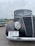 A vintage black truck with Wisconsin license plate A37 37 displayed on a wet pavement. The truck features a large round headlight and a prominent grille design. Another vehicle, possibly another classic car, is partially visible in the background, set against a cloudy sky and a grassy field.