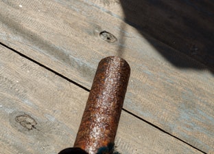 A rusted metal pipe is placed on a wooden surface with visible texture and knots. Sunlight casts a shadow, highlighting the corrosion on the pipe.