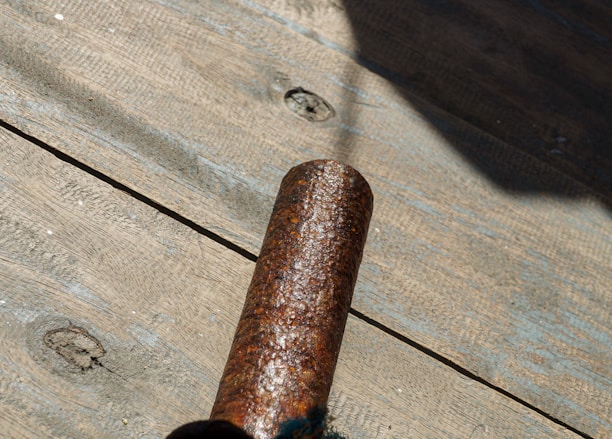 A rusted metal pipe is placed on a wooden surface with visible texture and knots. Sunlight casts a shadow, highlighting the corrosion on the pipe.