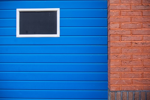 A section of a building featuring a blue paneled wall with a small rectangular window bordered in white. Adjacent to the blue wall is a contrasting red brick wall.