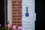 A decorative blue eye charm known as a Nazar amulet is hanging on a section of a white brick wall. To the left, there is red brickwork contrasting against the white. In the foreground, blurred pink flowers add a splash of color, providing a sense of depth to the photograph.