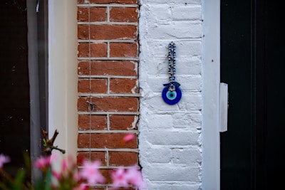 A decorative blue eye charm known as a Nazar amulet is hanging on a section of a white brick wall. To the left, there is red brickwork contrasting against the white. In the foreground, blurred pink flowers add a splash of color, providing a sense of depth to the photograph.