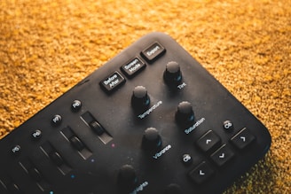 A black control panel with various knobs and buttons labeled with functions such as Temperature, Tint, Saturation, and Vibrance. The panel has smooth, round knobs and slider controls, with a textured surface. The background is a warm, textured gold color, possibly a carpet or fabric.