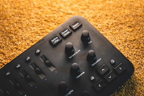 A black control panel with various knobs and buttons labeled with functions such as Temperature, Tint, Saturation, and Vibrance. The panel has smooth, round knobs and slider controls, with a textured surface. The background is a warm, textured gold color, possibly a carpet or fabric.