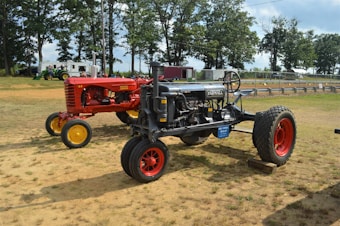 Two vintage tractors are parked on a grassy field, surrounded by trees and a few vehicles in the background. The tractor on the left is red with yellow wheels and labeled 'Massey Harris', while the tractor on the right is dark gray with red wheels and labeled 'Farmall'. The scene appears to be part of a show or exhibition, with a serene rural setting.