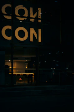 Dimly lit interior space with a reflective surface showing the word 'CON'. The lighting creates a warm atmosphere with a focus on a modern interior design visible through a window.