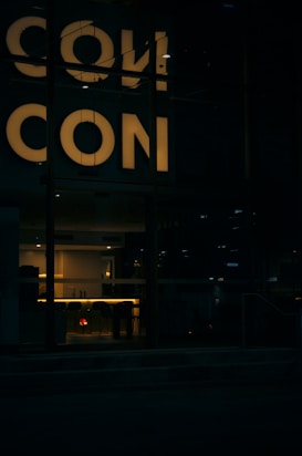 Dimly lit interior space with a reflective surface showing the word 'CON'. The lighting creates a warm atmosphere with a focus on a modern interior design visible through a window.