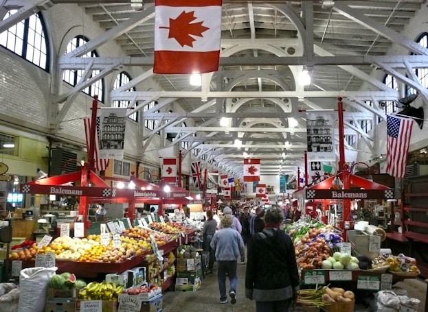 A bustling indoor market with a variety of fresh produce displayed on stalls. Multiple flags, including Canadian and American, hang from the ceiling. The market is filled with people walking and shopping.