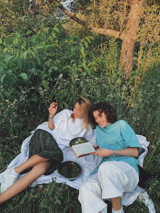 two women sitting on picnic blanket reading a book and smiling