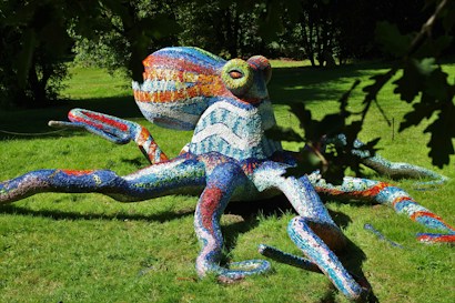 A colorful mosaic sculpture of an octopus is situated on a green lawn, surrounded by trees. The sculpture is made up of various colored tiles, including blue, red, orange, and white, giving it a vibrant and artistic appearance.