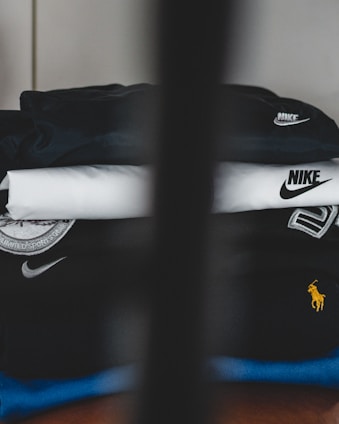 A neatly stacked pile of clothing with visible brand logos, including Nike and a horse logo. The clothes are mainly in dark and neutral colors, with a prominent black piece featuring a large white swoosh. A subtle vertical obstruction divides the image, adding a sense of depth.