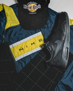 A Hard Rock Cafe London cap, a Nike sports jacket with a yellow, white, and blue color scheme, and a black Nike shoe are arranged. The items are laid out on a flat surface, showcasing a mix of casual and sporty attire.