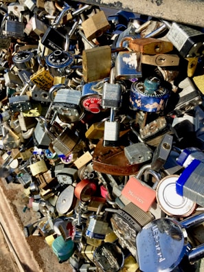 A cluster of various types of padlocks attached to a metal rail, creating a dense and colorful pattern. Some locks are rusty, while others are shiny and new, with different designs, brands, and colors.