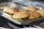 Several hamburgers arranged on a metal tray, each containing a patty with lettuce inside soft buns. The surface below the tray appears to be part of a kitchen setting.