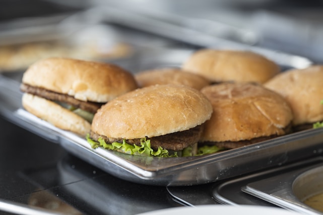 Several hamburgers arranged on a metal tray, each containing a patty with lettuce inside soft buns. The surface below the tray appears to be part of a kitchen setting.