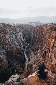 A dramatic canyon landscape with steep rocky cliffs stretching towards the horizon. A narrow river cuts through the base of the canyon, and a bridge spans across the top, connecting both sides. Sparse vegetation is visible on the cliff edges.