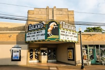 A vintage brick building featuring an old-fashioned theater marquee with the name 'Island' in bold letters. Movie titles such as 'Misty' and 'Dora: Lost City of Gold' are displayed. The building's facade includes decorative elements, glass doors, and poster displays. Nearby, small shops are visible, and overhead power lines cross the scene.