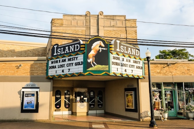 A vintage brick building featuring an old-fashioned theater marquee with the name 'Island' in bold letters. Movie titles such as 'Misty' and 'Dora: Lost City of Gold' are displayed. The building's facade includes decorative elements, glass doors, and poster displays. Nearby, small shops are visible, and overhead power lines cross the scene.